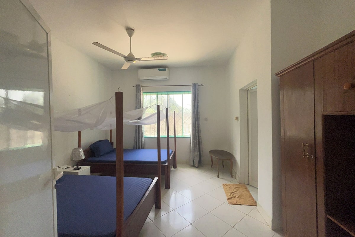 A bedroom with two single beds, a ceiling fan, an air conditioning unit, a window with curtains, and a wardrobe.