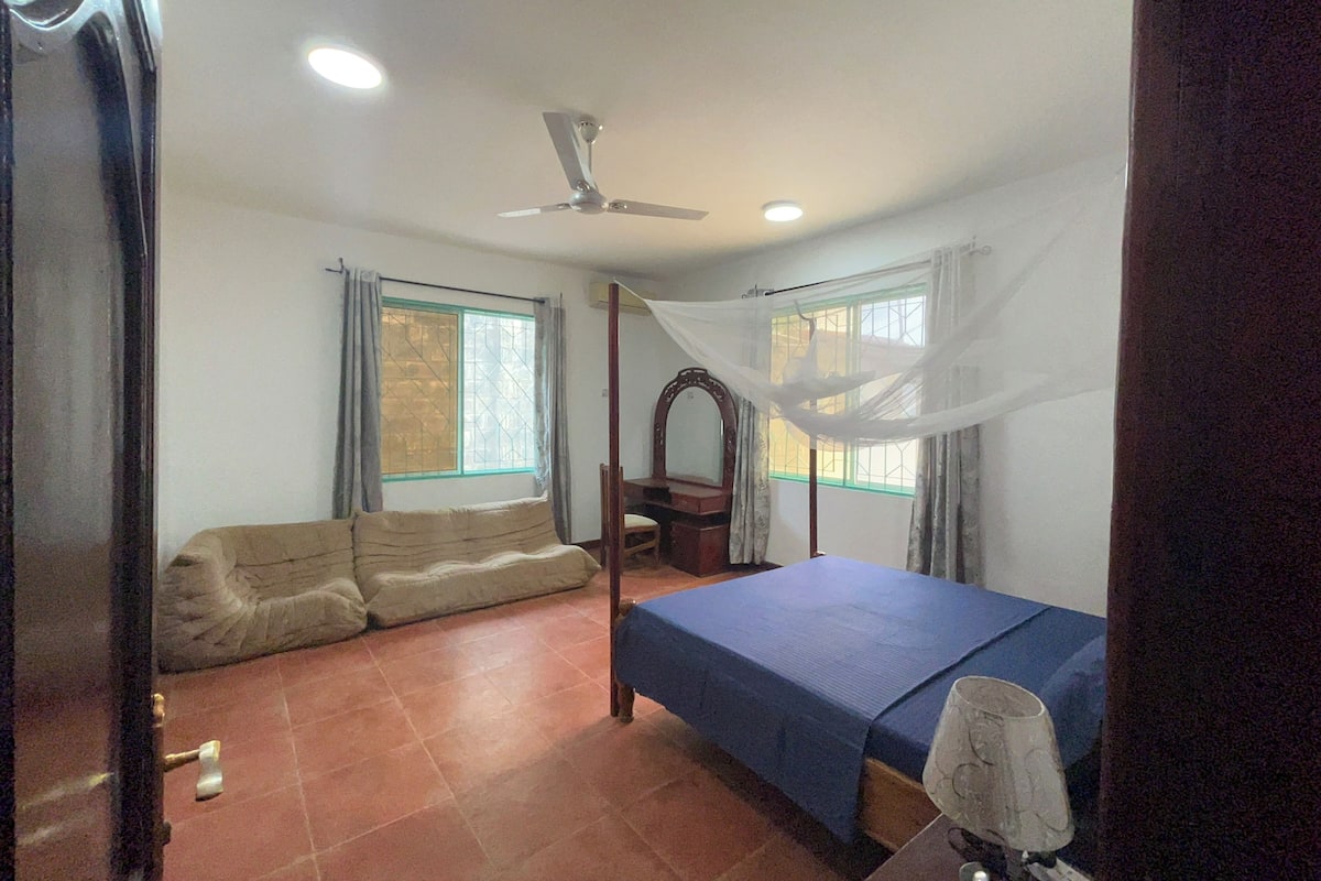 A bedroom with a bed, a sofa, a ceiling fan, and two windows with curtains.