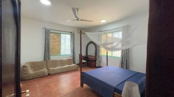 Un dormitorio con cama, sofá, ventilador de techo y dos ventanas con cortinas.