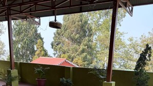 A covered outdoor seating area with wicker chairs and a glass-top table, surrounded by potted plants and a view of trees and a building with a red roof. - K2view Retreat (Darjeeling)