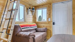 A wooden cabin interior with a brown leather sofa, a ladder, and a window with a view of a tree. - Cute Couple's Tiny Home, 8 Mi to Downtown Helena! (East Helena)
