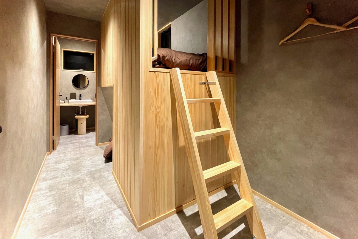A room with a bunk bed, a wooden ladder, a small bathroom, and a wooden wardrobe.