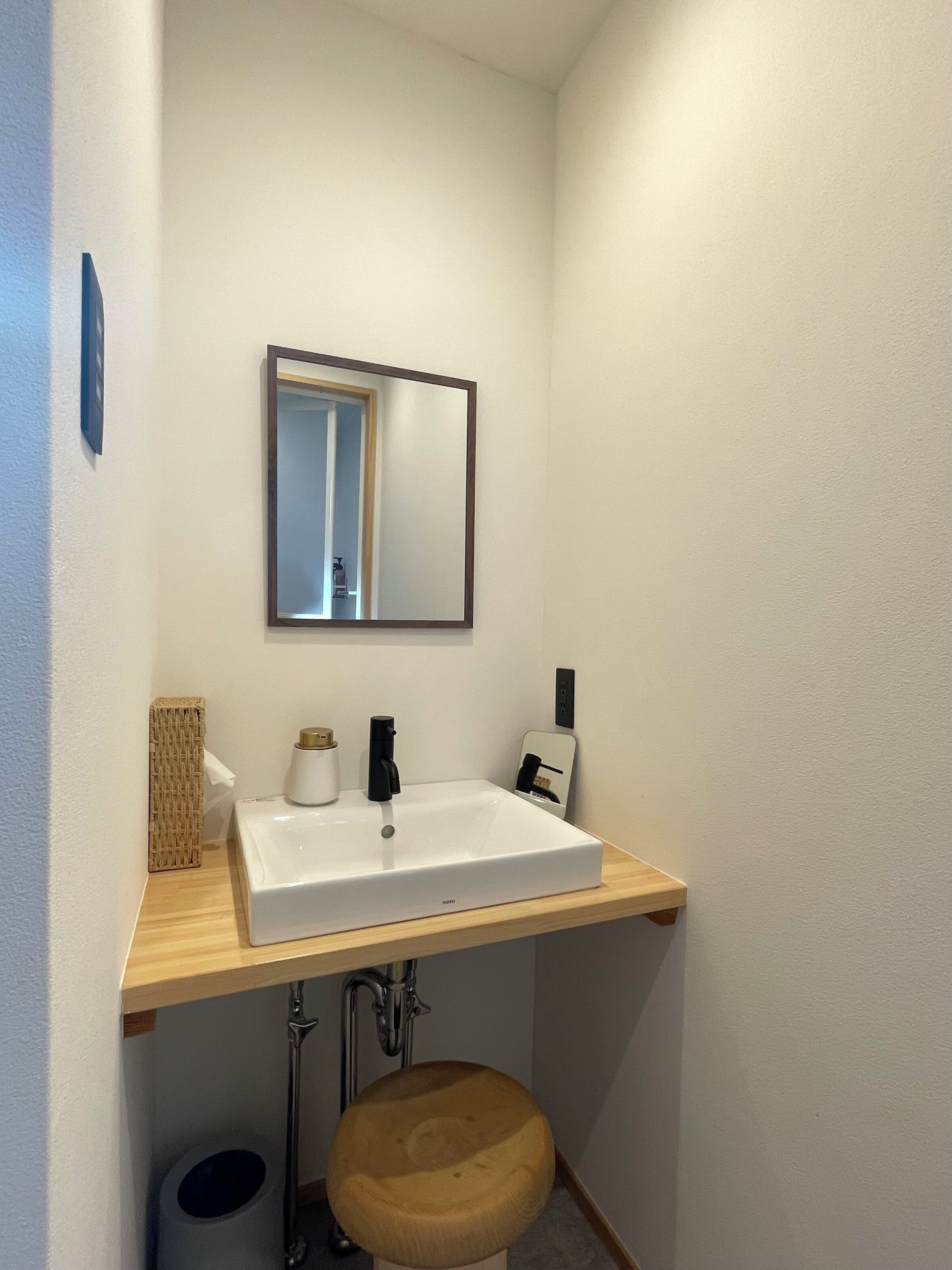 Ein Badezimmer mit einem weißen Waschbecken, einem Spiegel, einem Holzschemel und einem Handtuchspender.