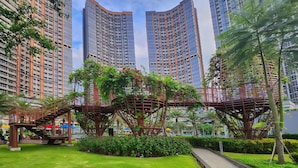 Ein moderner Stadtpark mit einer Fußgängerbrücke, üppigem Grün und hoch aufragenden Gebäuden im Hintergrund.