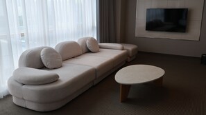A modern living room with a beige sectional sofa, a round coffee table, and a flat-screen TV mounted on the wall. - Centum Suite Hotel (Busan)