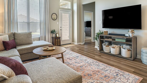 Ein Wohnzimmer mit einer Couch, einem Couchtisch und einem Fernseher. Es gibt einen Holzboden, einen gemusterten Teppich und ein Bücherregal mit dekorativen Gegenständen.