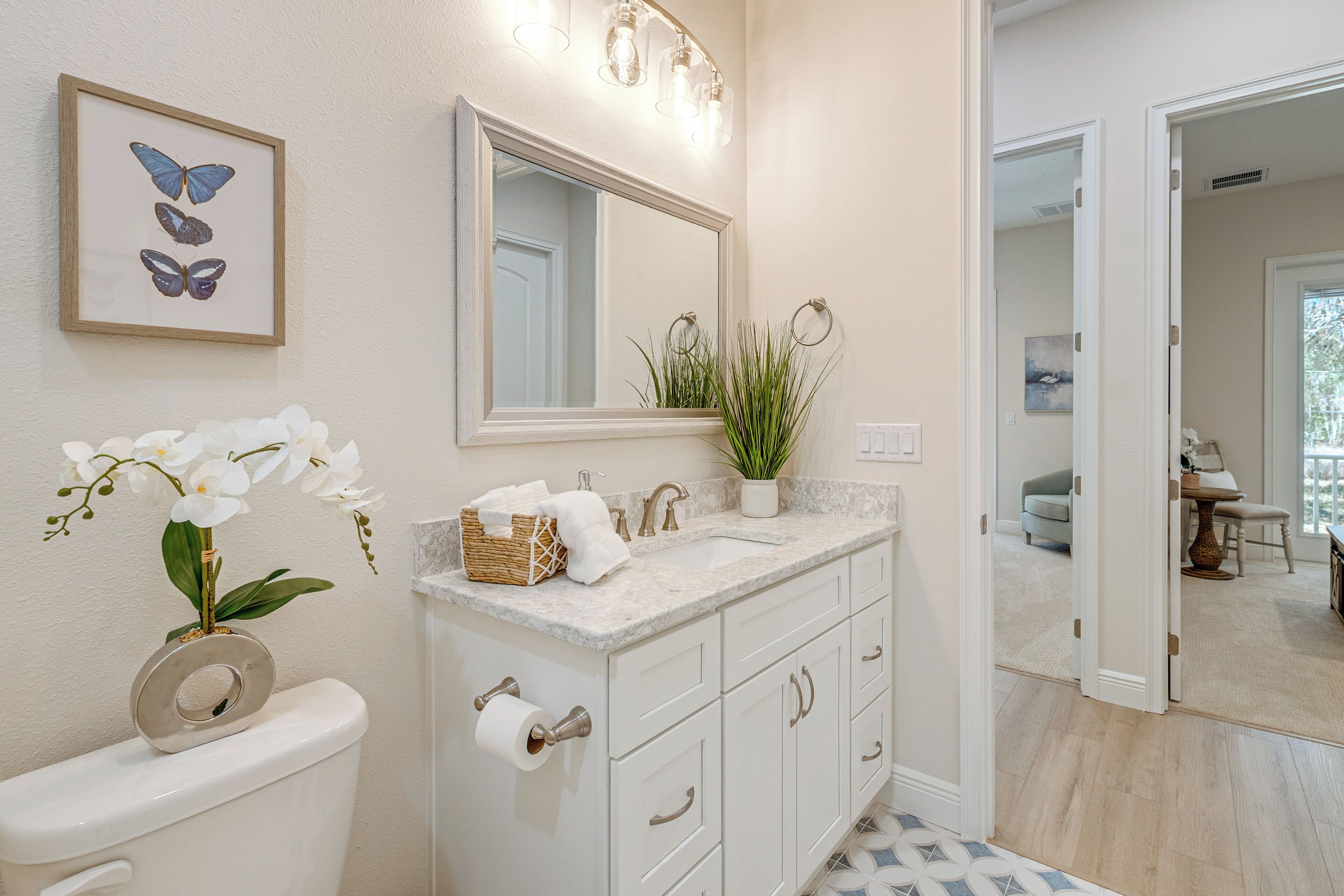 Un baño con inodoro, lavabo y espejo. Hay una obra de arte con una mariposa enmarcada sobre el espejo y una planta en maceta en la encimera del lavabo.