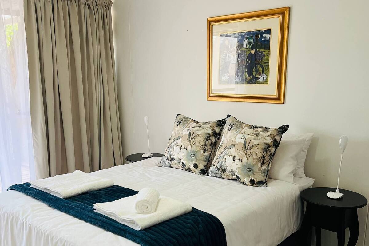 Una cama bien hecha con ropa de cama blanca y azul, dos almohadas estampadas con flores, un cuadro en la pared y una mesita de noche negra pequeña.