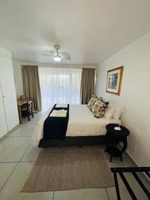 A bedroom with a bed, a desk, a chair, a ceiling fan, a window with curtains, and a framed picture on the wall.