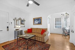 A living room with a striped sofa, a glass coffee table, a patterned rug, a ceiling fan, and a painting on the wall. - Pet Friendly Midtown Mobile Condo (Mobile)