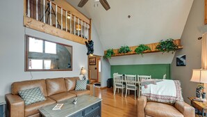 A spacious living room with a high ceiling, a wooden staircase, a dining area with a table and chairs, and a green accent wall.