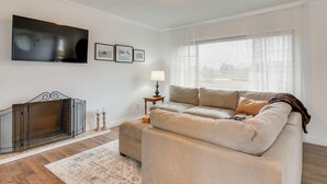 A living room with a large flat-screen TV mounted on the wall, a beige sofa, a small side table with a lamp, and a fireplace with a glass enclosure. - Cozy Mtn-View Retreat w/ Yard: Near Hiking & Golf! (Ferron)
