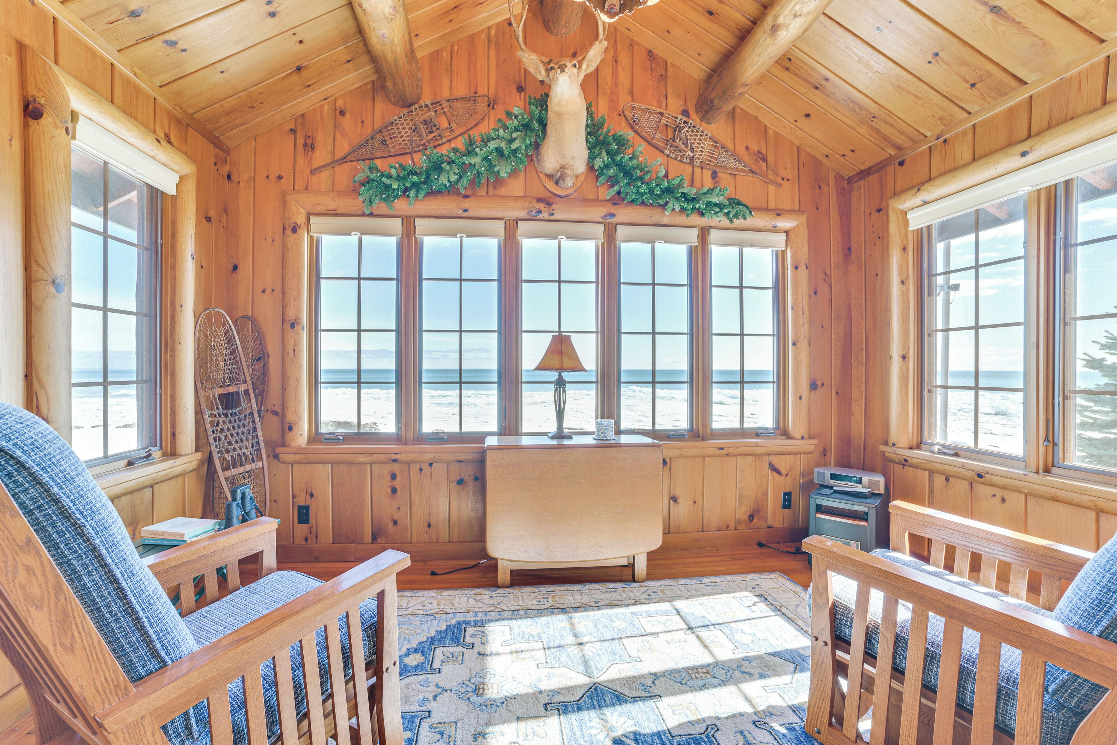 A cozy cabin with wooden walls, a large window with a view of the beach, a fireplace, and a ceiling adorned with antlers.