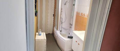 Een smalle badkamer met een bad, een douche en een klein kastje.