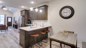 Een moderne keuken met donkere kasten, een centraal eiland met krukken, een hangend horloge en een eetgedeelte met een houten tafel.