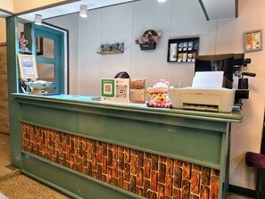 Una taquilla amb mostrador verd, una paret decorativa amb relleu de maó, un cartell amb codi QR, una màquina de cafè i un panell informatiu amb el menú.