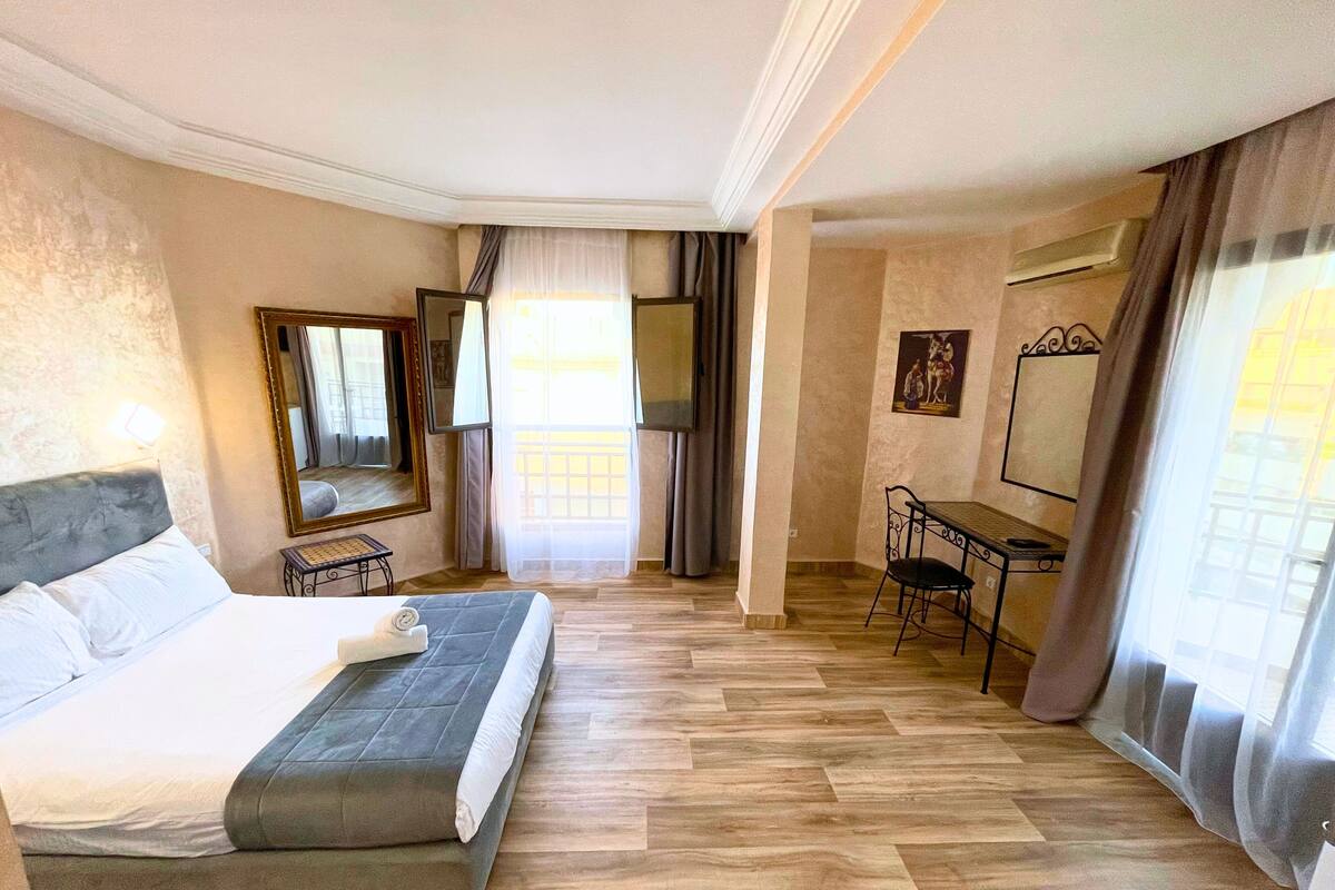 A hotel room with a large bed, a desk, and a chair. There is a window with curtains, a mirror, and a painting on the wall.