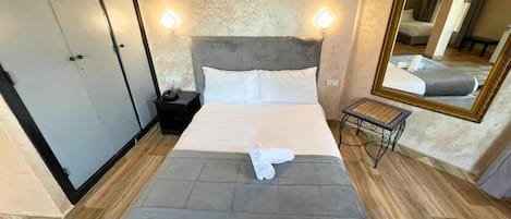 Una cama bien hecha con cabecera gris, dos lámparas de noche y una mesita con una silla.