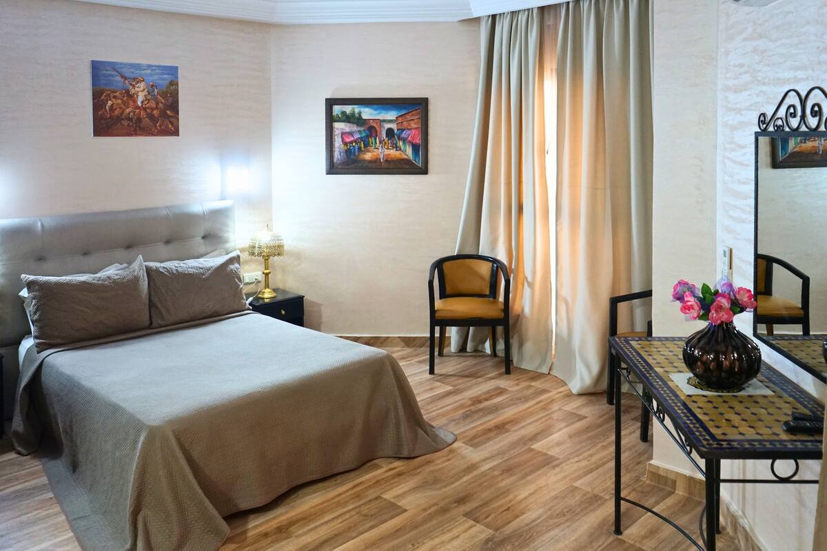 A bedroom with a bed, a chair, a table with a vase, and two paintings on the wall.