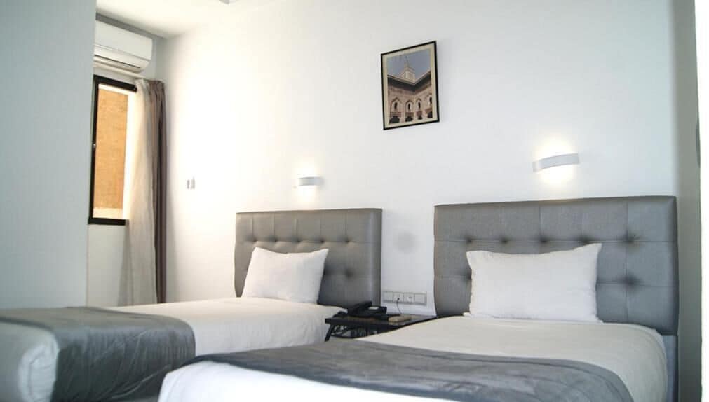 Een hotelkamer met twee bedden, elk met een grijze hoofdbord en witte beddengoed. Boven de bedden hangt een ingelijste foto aan de muur.