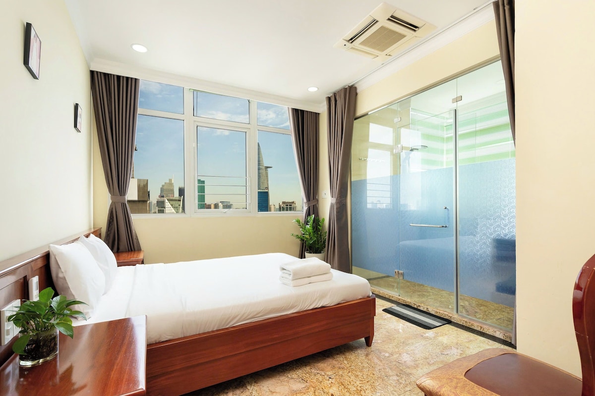 A hotel room with a bed, a desk, a chair, a potted plant, and a sliding glass door leading to a balcony.