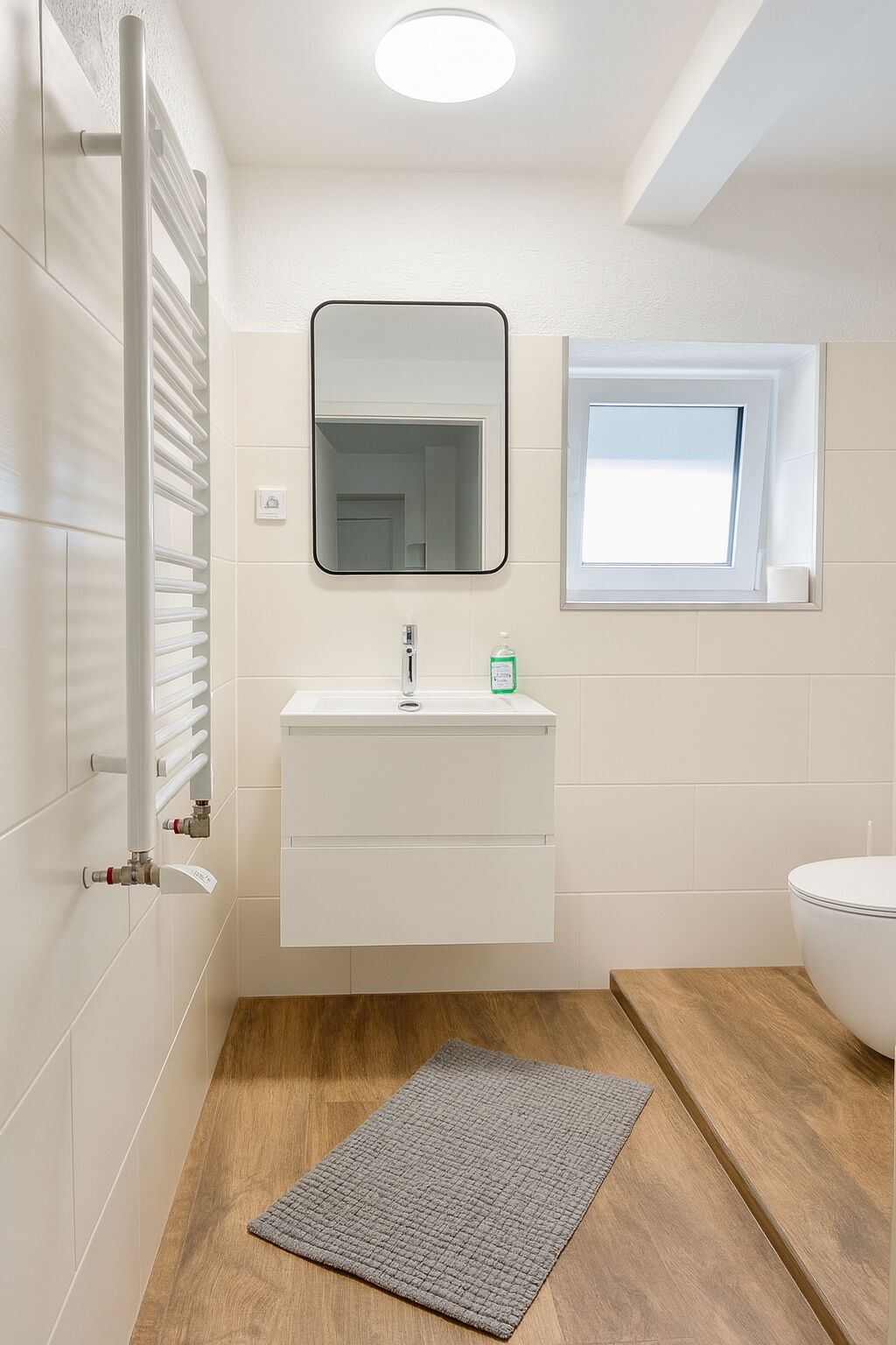 Een badkamer met een wit wastafelmeubel, een spiegel, een raam en een houten vloer.