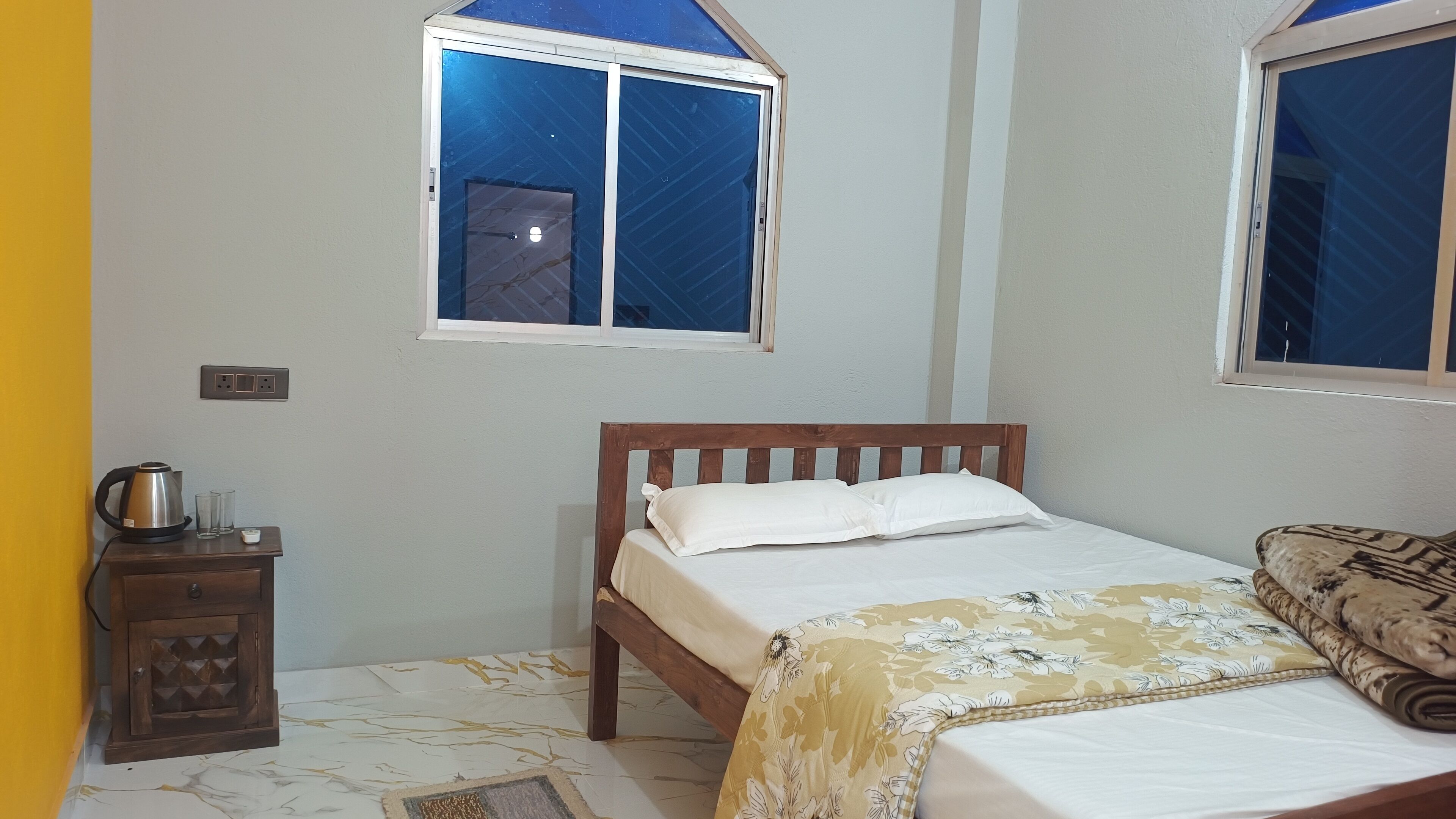 Un dormitorio con cama, mesita de noche, ventana y enchufe.