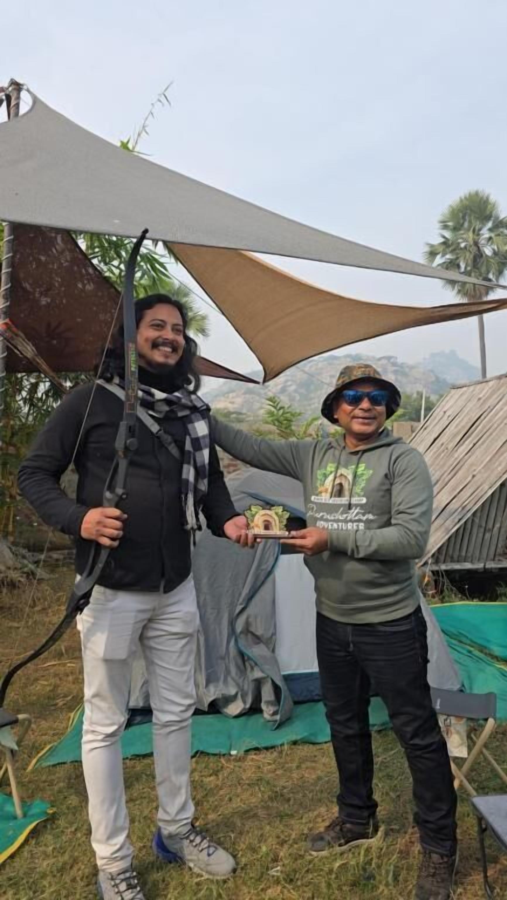 Due persone che stringono un trofeo, in piedi sotto una grande tettoia in un ambiente esterno con una tenda e palme sullo sfondo.