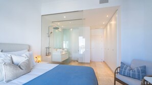 A modern bedroom with a large bed, a sofa, and a bathroom visible through a glass door. - Modern Luxurious 3-Bedroom Residence with Resort Amenities (Benahavís)