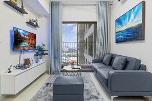 A modern living room with a flat-screen TV, a grey sofa, a white console, and a balcony with a view. - MASTERI Premier Residence By An Gia Hospitality (Ho Chi Minh City)