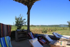 A covered outdoor seating area with lounge chairs and a wooden table, overlooking a vineyard.