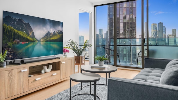 A modern living room with a large TV, a sofa, a coffee table, and a view of a cityscape through large windows. - Stunning 2-bed Apartment With City Views (Melbourne)