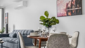A modern living room with a grey sofa, a dining table with chairs, a large plant, and a painting of a forest. - Stunning 2-bed Apartment With City Views (Melbourne)
