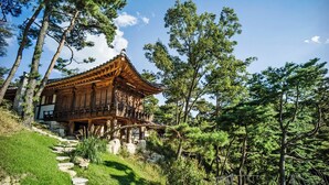 A traditional wooden building with a sloped roof, surrounded by trees and a stone pathway. - L'art de la vie Art & Resorts (Icheon)
