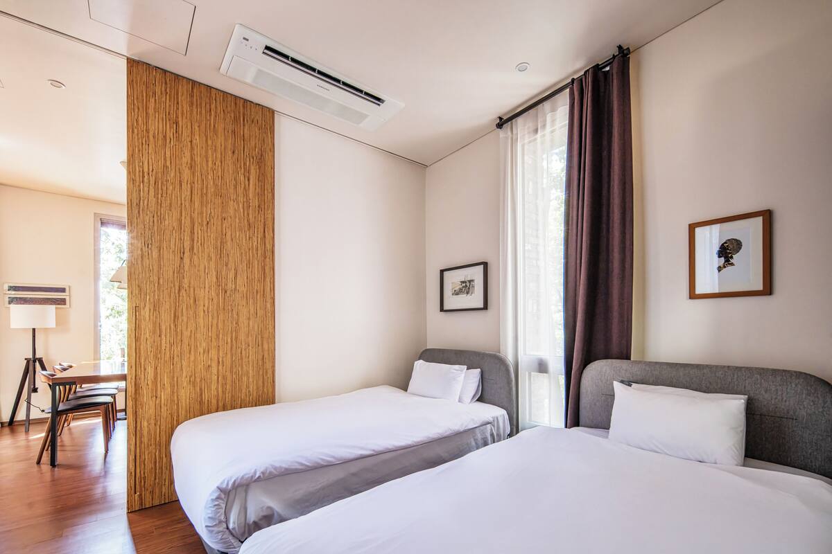 A hotel room with two beds, a desk, a chair, a framed picture, and an air conditioning unit.