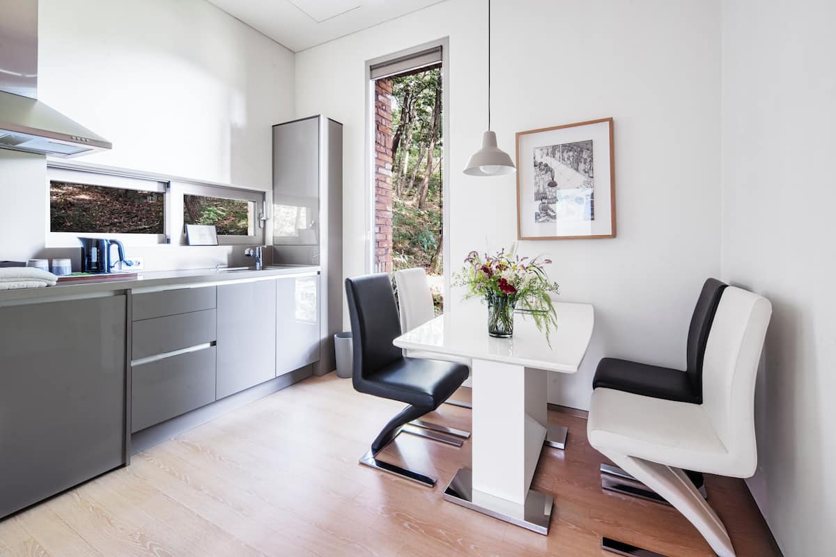 A modern kitchen with a dining area, featuring a white table, black chairs, and a view of trees through a window.