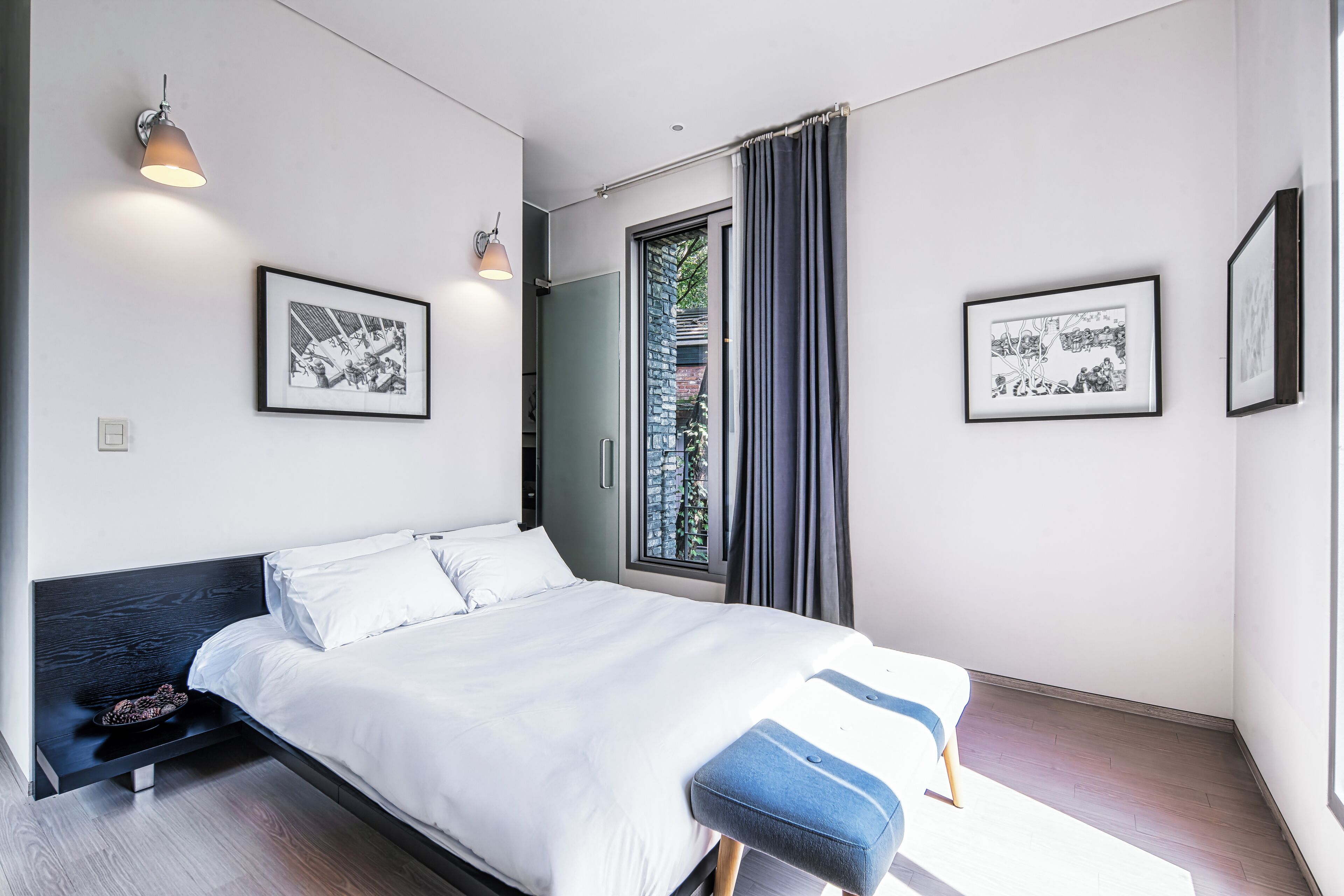 Une chambre à coucher moderne avec un lit, un banc, une lampe fixée au mur et des œuvres d’art encadrées.