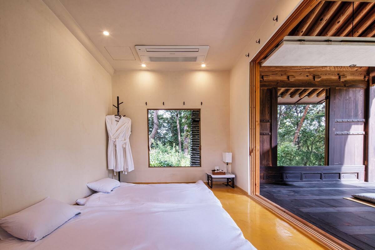 A bedroom with a large bed, a window with a view of trees, a small table with a lamp, and a wooden ceiling.