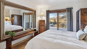 Ein Schlafzimmer mit einem großen Bett, einer hölzernen Bank, einem Spiegel, einer Lampe und Blick auf eine verschneite Landschaft durch ein Fenster.