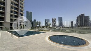 Un edificio residencial moderno con zona de piscina en la azotea, que cuenta con una piscina rectangular grande y una piscina circular más pequeña, rodeadas por un patio amplio.
