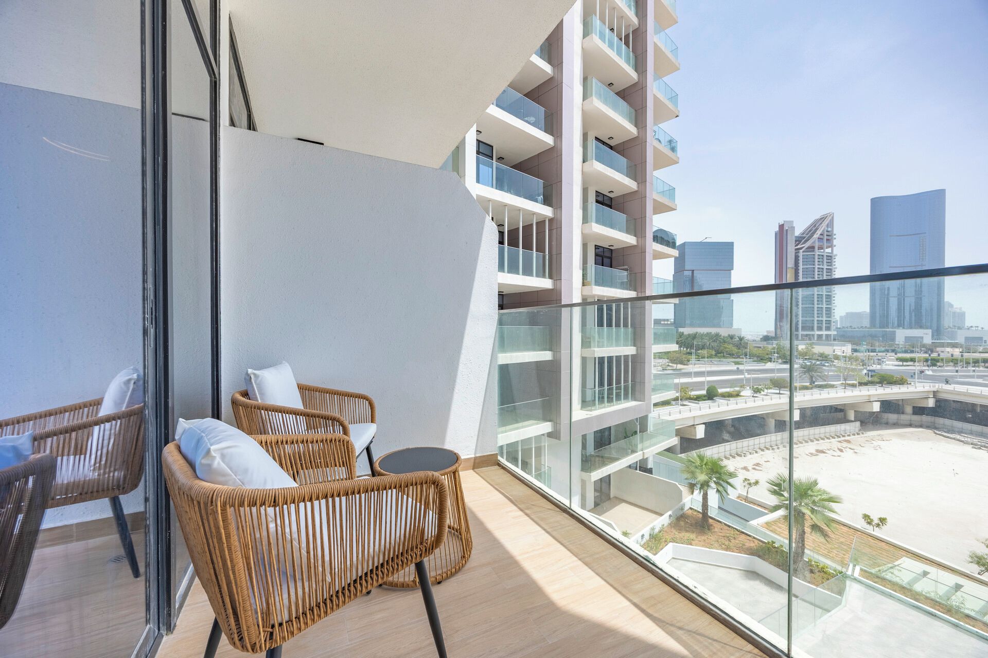 A balcony with wicker furniture, a glass railing, and a view of a modern cityscape and beach.
