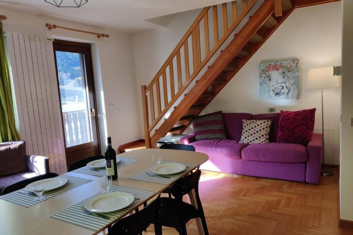 A dining area with a wooden table set for two, a purple sofa, and a staircase leading to an upper level.