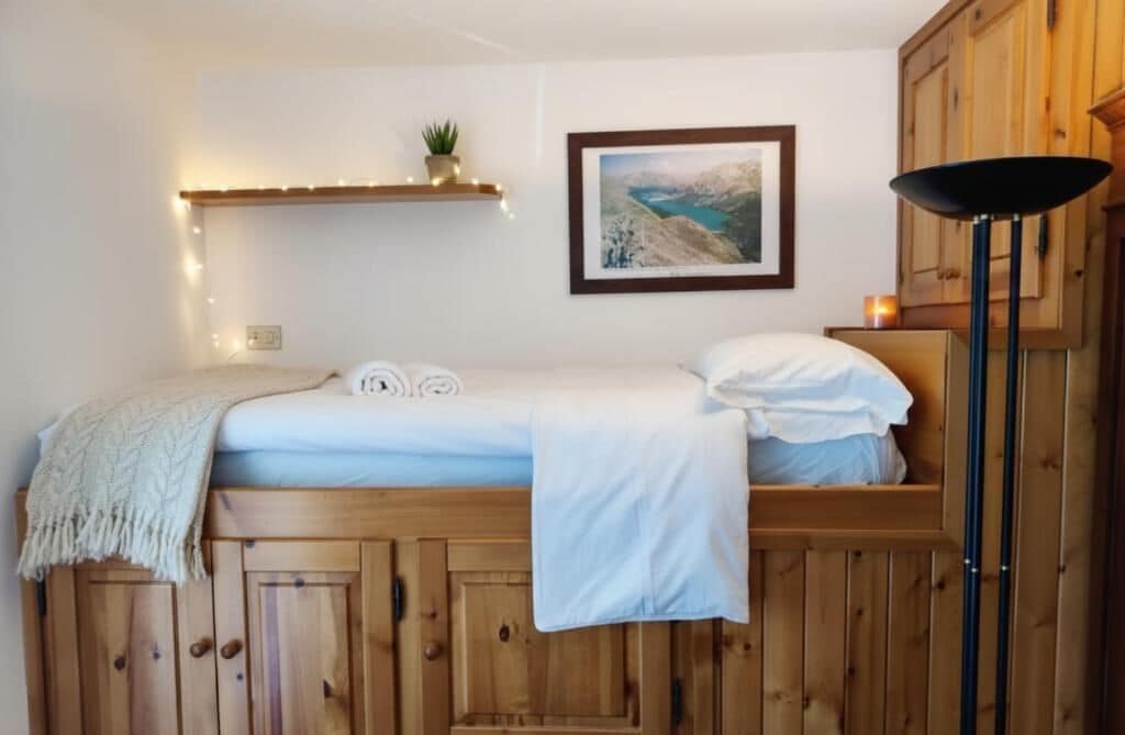 A wooden bed with a white blanket, a framed picture on the wall, and a shelf with lights and a plant.