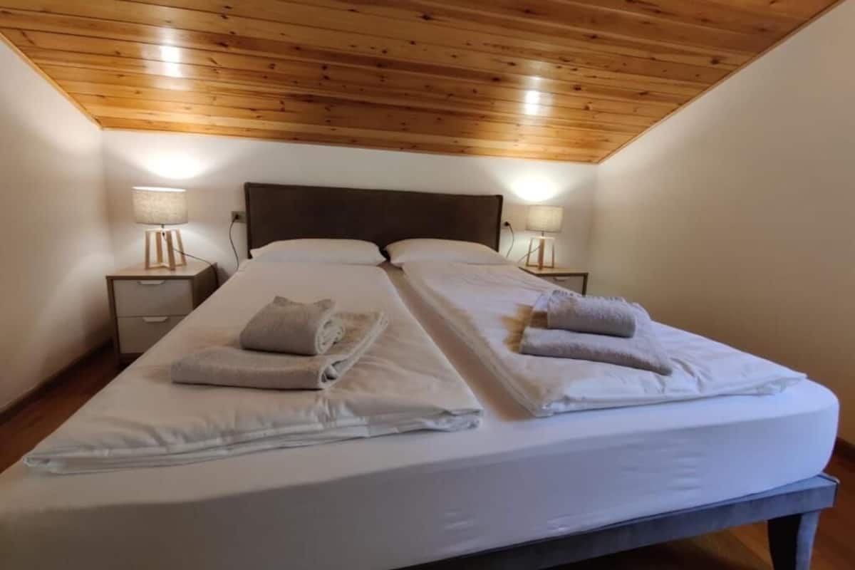 A double bed with white linens and towels, wooden ceiling, two bedside tables with lamps, and a nightstand.
