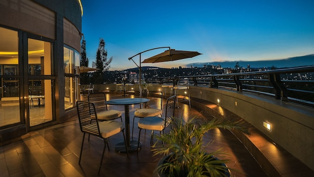 Terraço no telhado com mesas e cadeiras, um vaso de planta e vista para o horizonte urbano ao anoitecer.