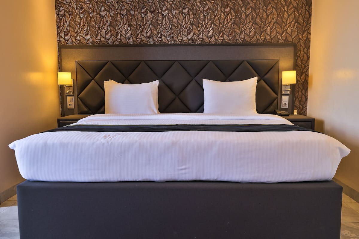 Une chambre d’hôtel avec un grand lit, deux lampes de chevet, une tête de lit sombre et un papier peint à motifs.
