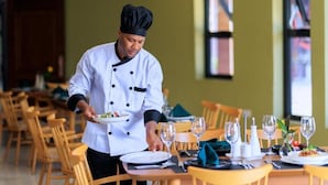 Um chefe de cozinha vestindo uniforme branco serve um prato para uma mesa posta com taças de vinho e pratos.
