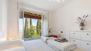 Ein Schlafzimmer mit Bett, Nachttisch, Kommode, einer Vase mit getrockneten Blumen und einem Balkon mit Blick auf Bäume.