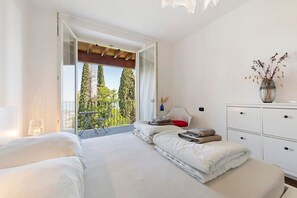 Ein Schlafzimmer mit Bett, Nachttisch, Kommode, einer Vase mit getrockneten Blumen und einem Balkon mit Blick auf Bäume.
