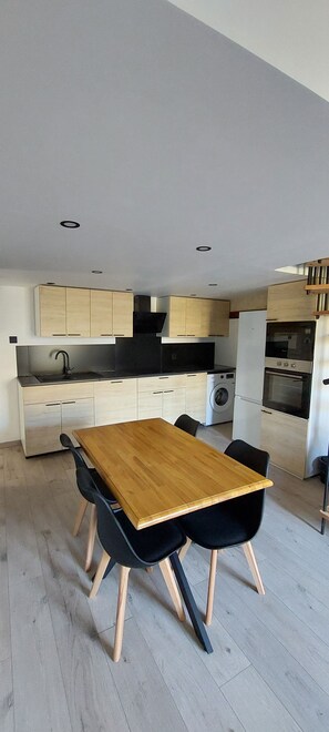 A modern kitchen with a wooden table and black chairs, light-colored cabinets, and built-in appliances. - Gite Rive Charente La Cabane (Bords)